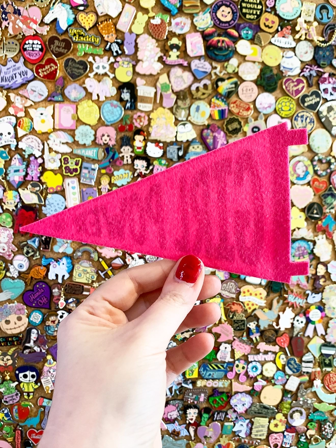 image shows a hand holding a bright pink felt pennant. only the back of the pennant shows, it is plain pink with the faint words from the print on the other side showing through.