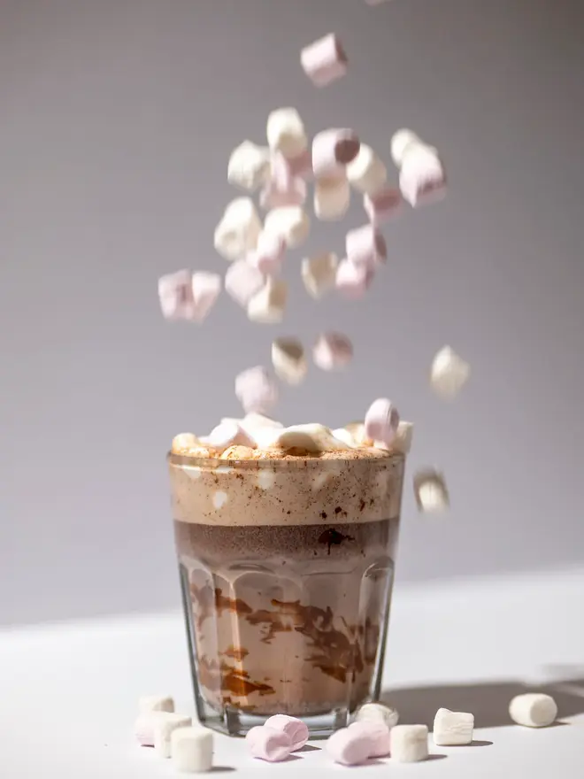 Cup of hot chocolate on a white table, mini marshmallows are being dropped in.  