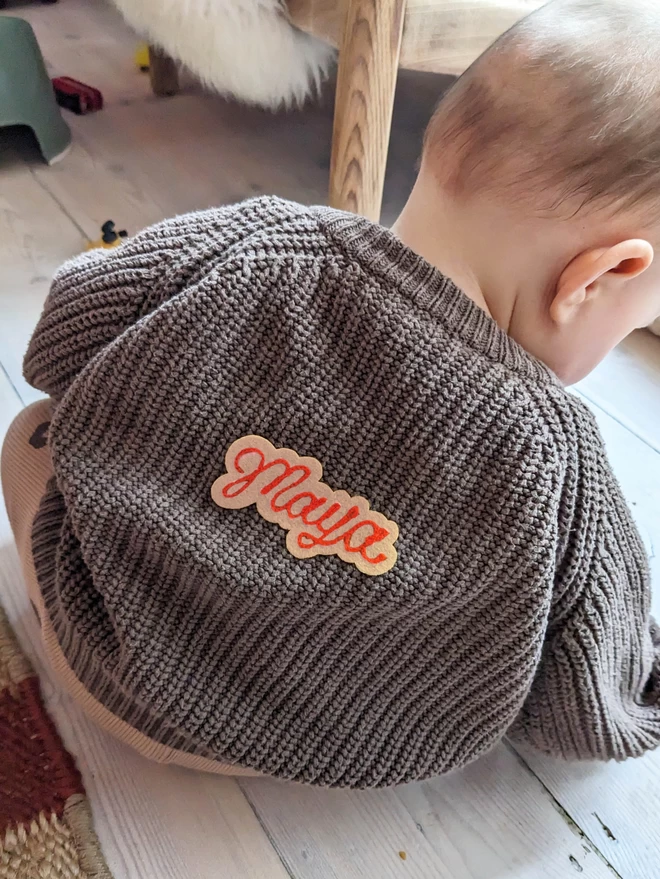 Ivory felt with red embroidered cursive lettering reading the name 'Maya' on the back of a baby girls cardigan as she sits on a white wooden floor