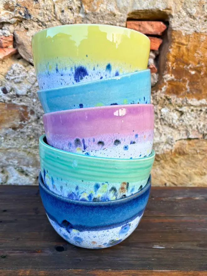 Shoreline Small Bowl, a group of colourful bowls stacked on top of each other on a wooden surface in front of a brick wall. 