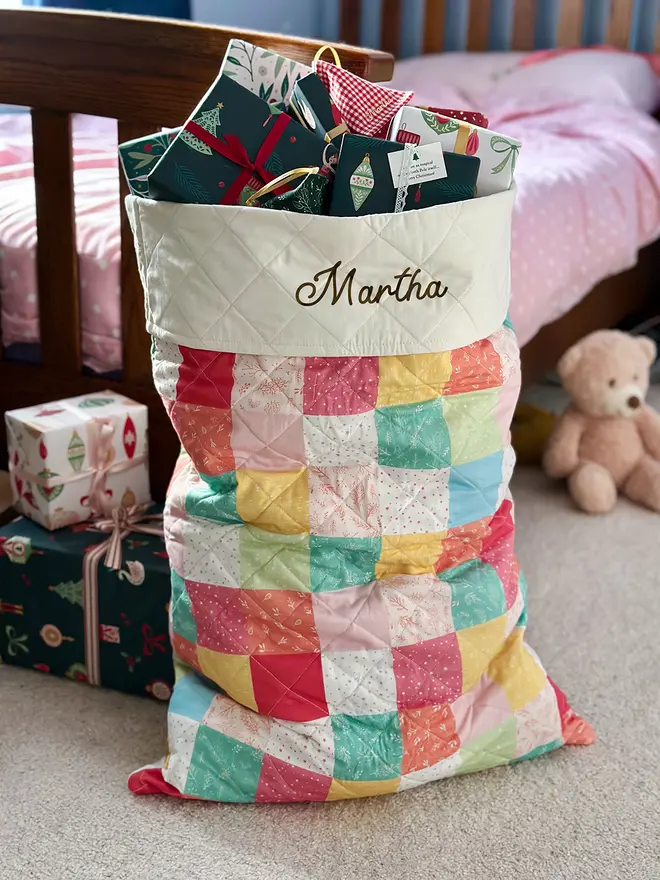 A handmade pastel patchwork Christmas sack stands beside a bed.