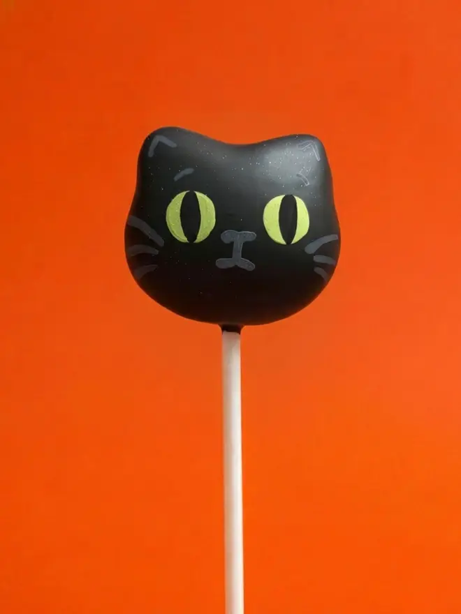 Black cat cake pop on a white stick against a bright orange background