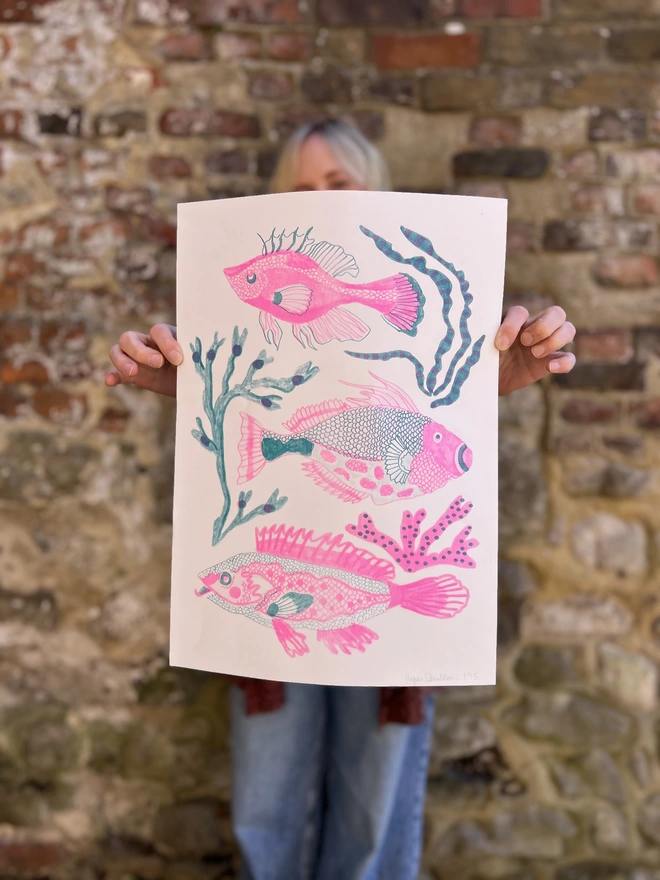 pink and blue risograph print of 3 fish and seaweed, held by person in front of brick wall