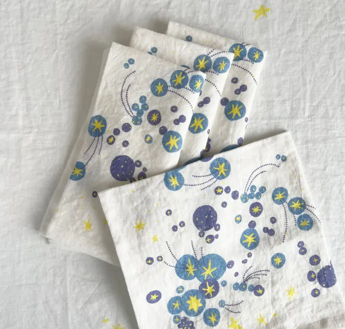 Solaris Napkins In White Or Blue. Set of four off-white cloth napkins with blue and yellow starburst and orbital patterns, stacked in a staggered arrangement.