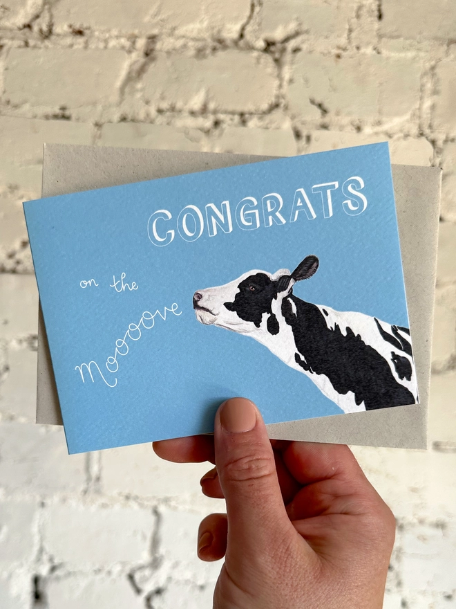 a sky blue card with an illustration of a black and white cow and the text "congrats on the moooove", with a light grey envelope