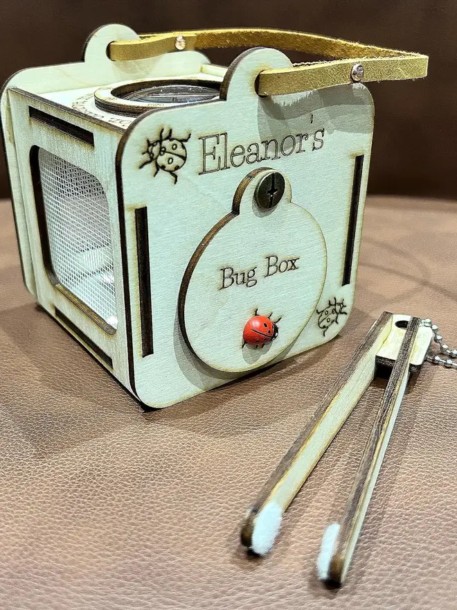 Baltic birch plywood bug observation box with clear viewing panels and a leather carrying handle displayed on table next to wooden insect-friendly tweezers. 
