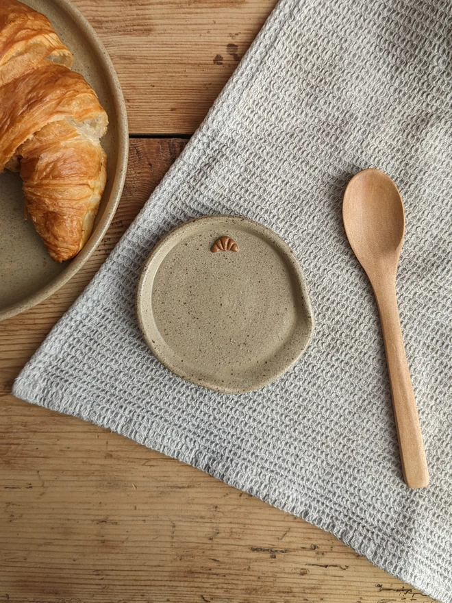 Stoneware spoon rest in biscuit glaze with a croissant design and a spoon beside it, on a wooden table.