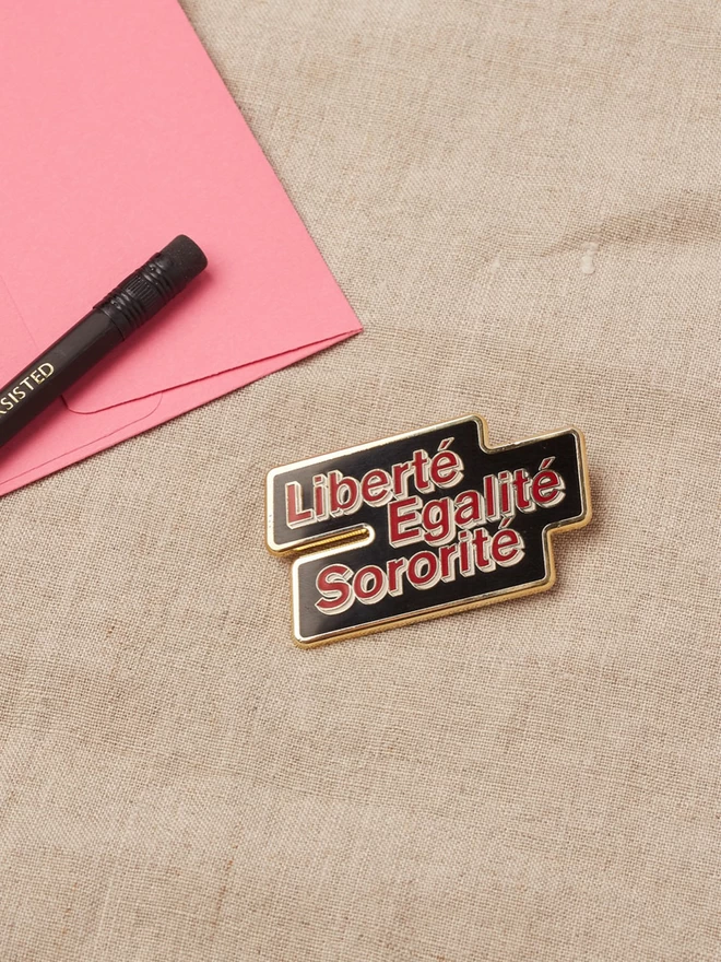 A black enamel pin with a gold edge with the words Liberté Egalité Sororité written in red with a white boarder.