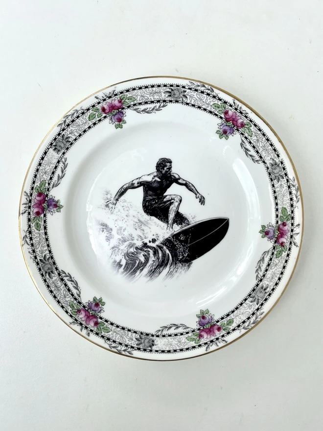 a vintage style image of a surfer riding a wave, 60s style, is printed in the centre of a vintage plate with an ornate edge