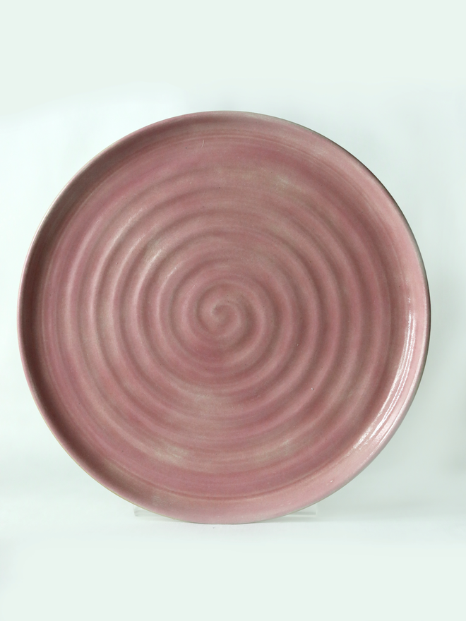 A handmade ceramic lunch plate in glossy blush pink glaze, photographed from above against a plain light grey background, showing a natural concentric swirl texture from the wheel-throwing process