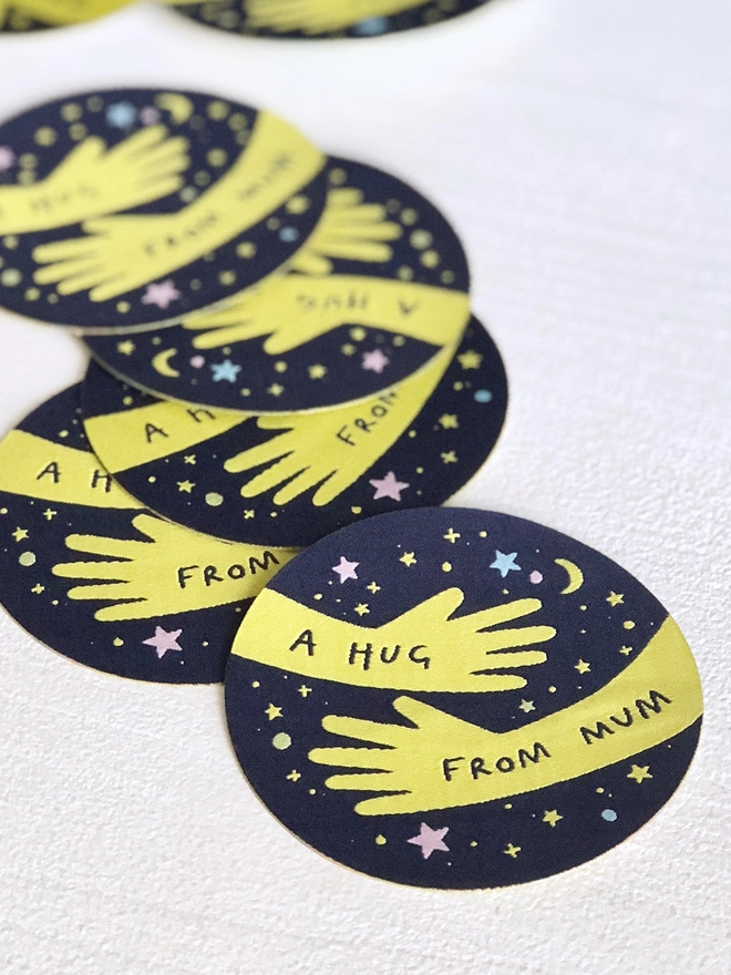 Several navy blue woven patches with a yellow hugging arm design and the words "A hug from Mum" are scattered on a white desk.
