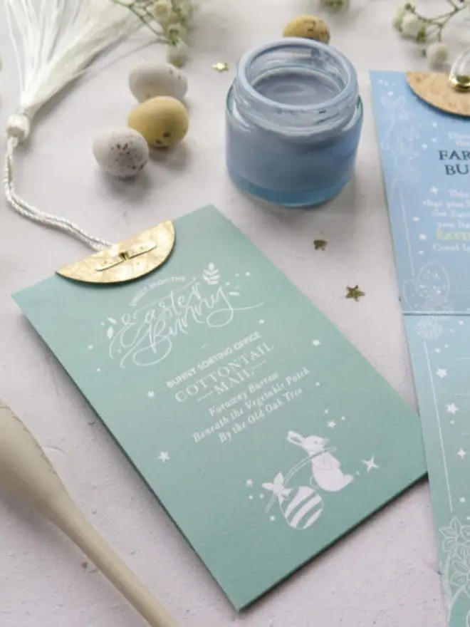A mint green "Cottontail Mail" card from the Bunny Sorting Office, with a white bunny pulling a striped egg. A blue paint jar, speckled mini eggs and a white tassel sit around it.