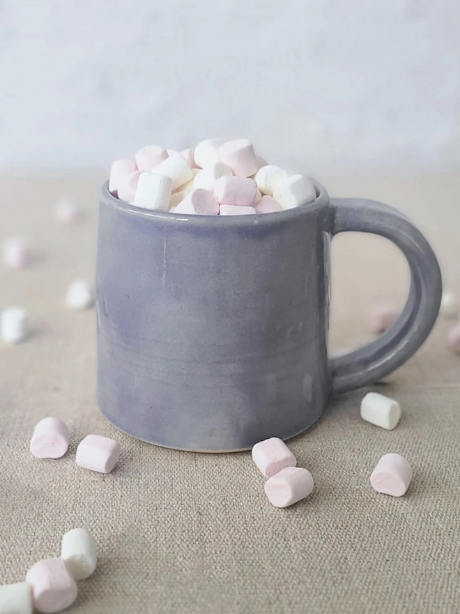 Ceramic Standard Mug, a colourful ceramic mug sitting on a plain surface against a grey backdrop. It is accompanied by marshmallows. 