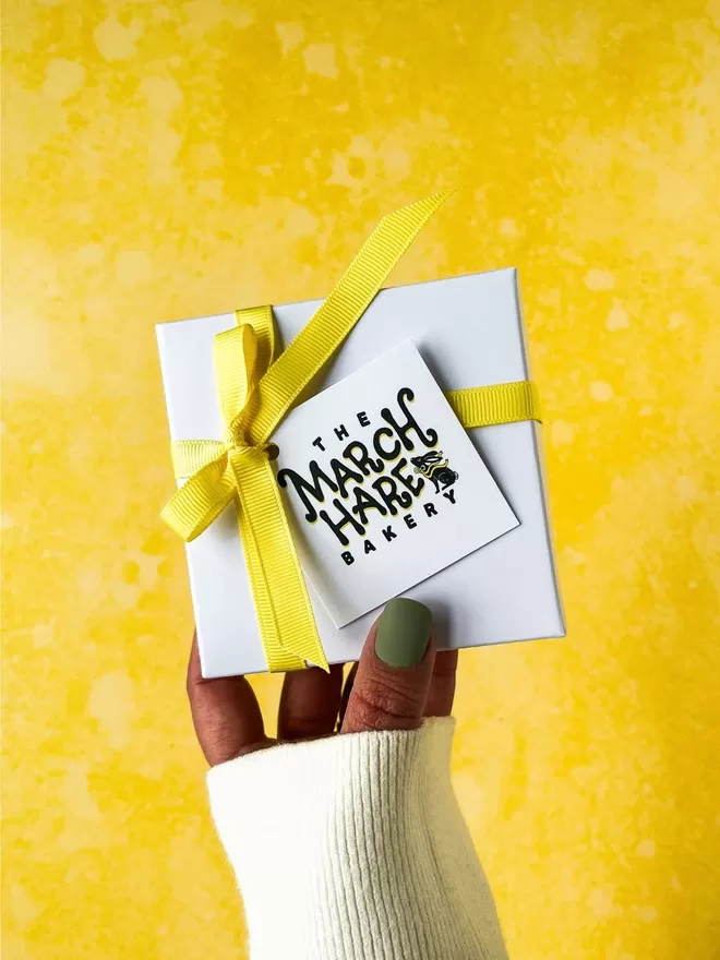 a female hand  with sage green nails holding up a white gift box with a yellow ribbon on it against a bright yellow background