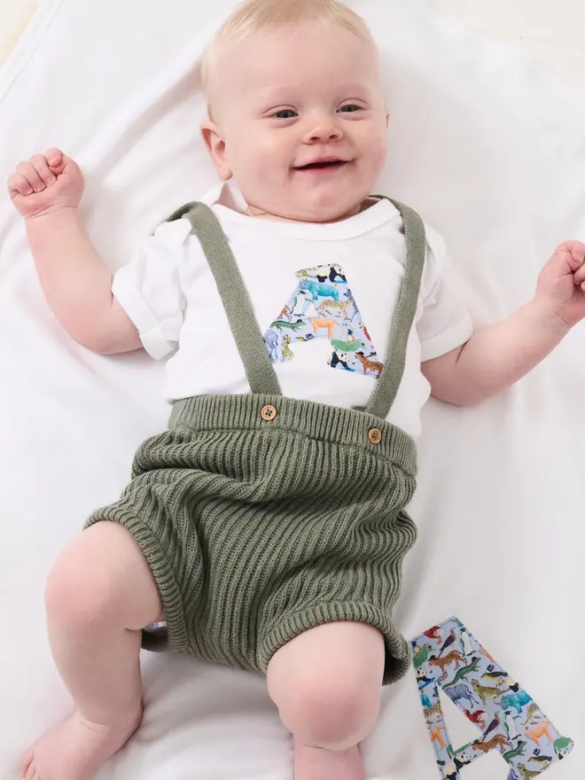 A smiling new baby wearing a bodysuit with his initial on the front cut from Liberty zoo animal print, laying on a  matching personalised blanket