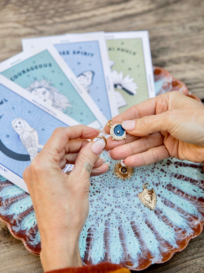 Older woman's hands holding a blue enamel crescent moon and star charm against an artisan blue plate