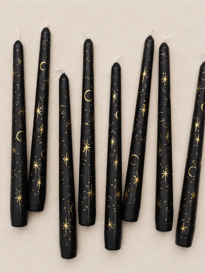Eight hand-painted black taper candles featuring gold celestial moons and stars, arranged at varying angles on a textured linen background.