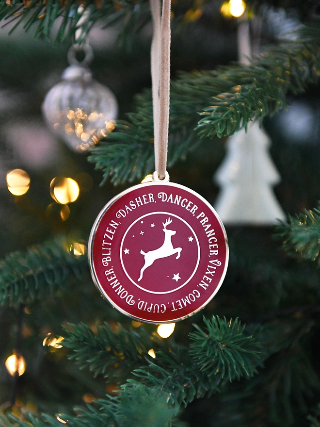 A deep red and gold enamel Christmas decoration, with the names of all eight reindeer surrounding a gold reindeer, is hanging on a Christmas tree.