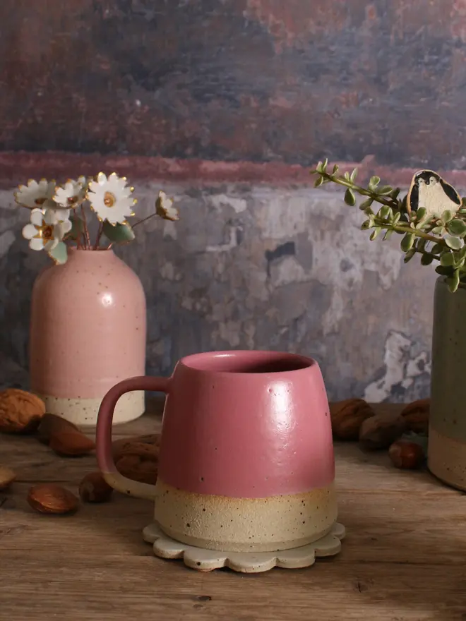 Ceramic Fuchsia mug sat on a ceramic coaster on a wooden surface. The background shows a pale pink ceramic vase holding a ceramic flower bouquet.  