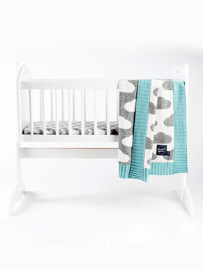 A product shot of a white rocking crib against a white background. A grey and white cloud pattern baby blanket with mint green ribbed trim is draped over the side of the crib. One side of the blanket is folded over to show the reverse colourway on the back.