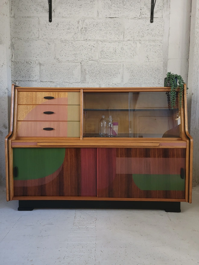 Mid-century teak sideboard with drawers and cupboard storage painted with translucent green and pink shapes.