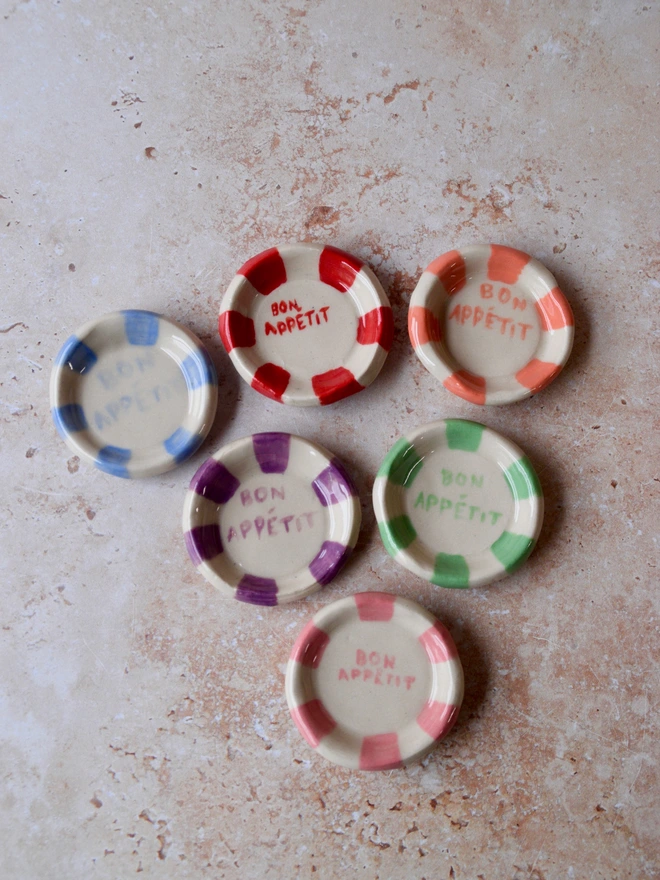 Handmade miniature ceramic dishes with hand-painted “Bon Appétit” text and colourful rim patterns in red, pink, purple, blue and green, arranged on a neutral background.
