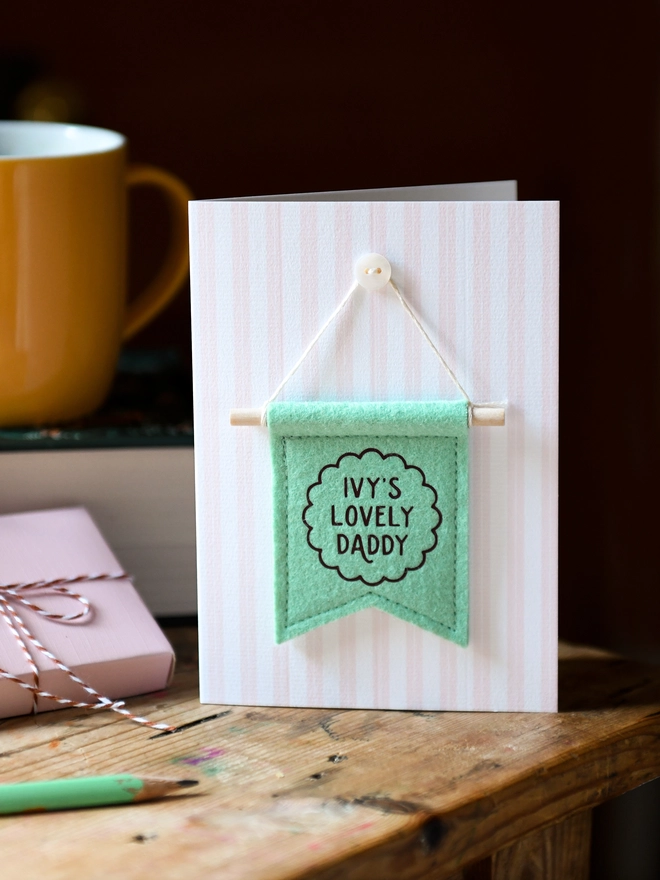 Handmade Father’s Day card with personalised green felt banner “Ivy's Lovely Daddy”, miniature hanging flag keepsake on wooden table. The flag is hung on the card from a tiny mother of pearl button. Beside the card is a book, a mug, a wrapped gift.