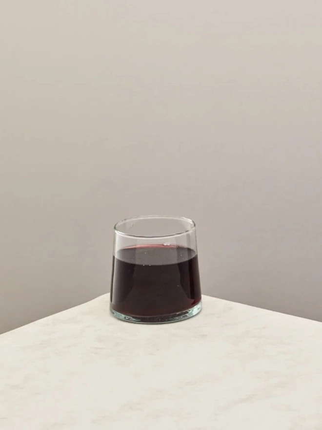Low Glasses (Set Of 1 Or 4), a low glass sitting on a white counter top against a plain backdrop. It is filled by an alcoholic drink. 
