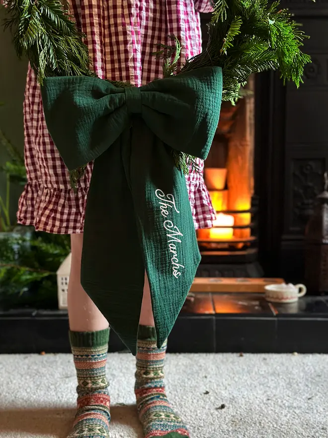 A handmade fabric Christmas bow decoration made from forest green cotton, with the personalisation 'The Marchs' on the right bow tail, is being held on a Christmas wreath by a child wearing a red gingham dress.