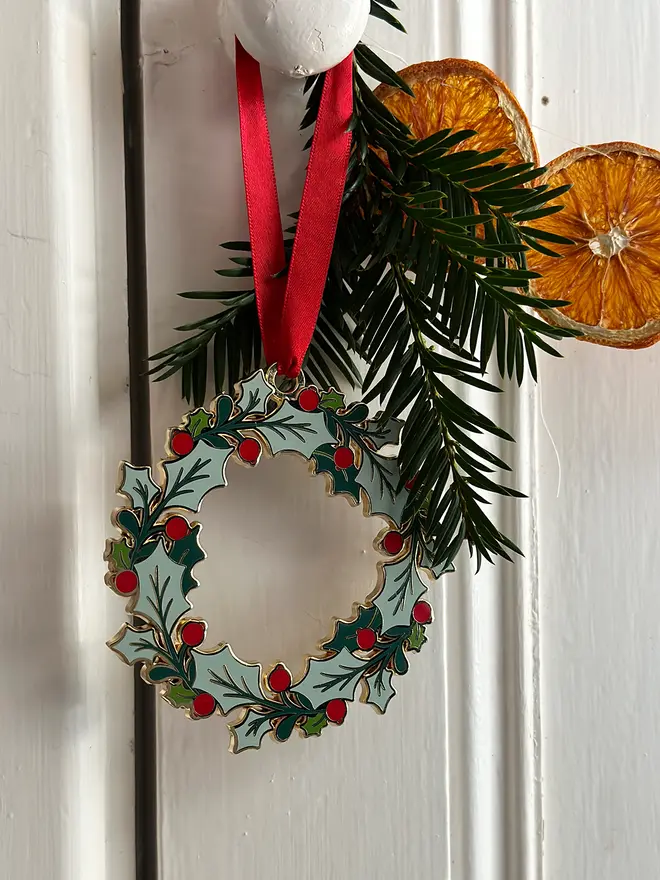 An heirloom Christmas wreath decoration with holly leaves and red berries hangs from a red ribbon on a white door handle with festive greenery and orange slice garland.