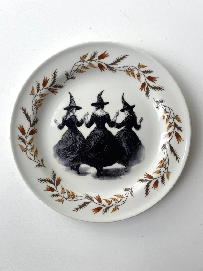 a coven of 3 witches dance hand in hand, the image is victorian in black and white and is printed directly onto an ornate vintage plate