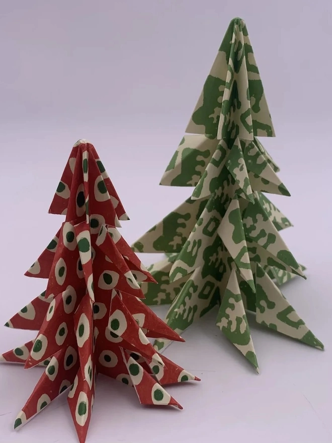 Christmas Tree Kits. Two origami Christmas trees folded from block-printed paper, one in red with green and cream spots and a taller one in green and cream leaf print. The layered paper trees make a handmade festive table or mantelpiece decoration.