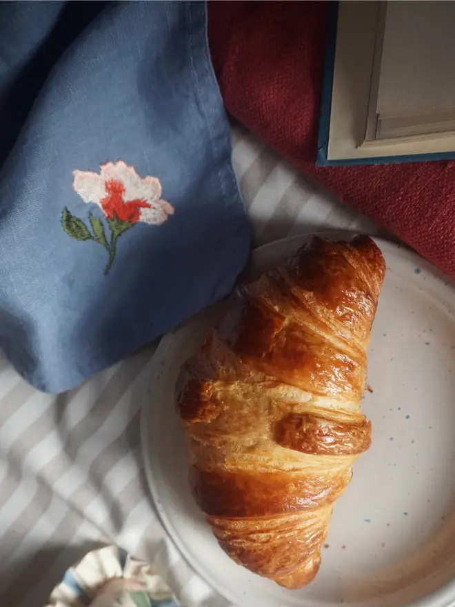 Natural linen napkin embroidered with blue and pink flowers, styled for breakfast with coordinating embroidered homeware.