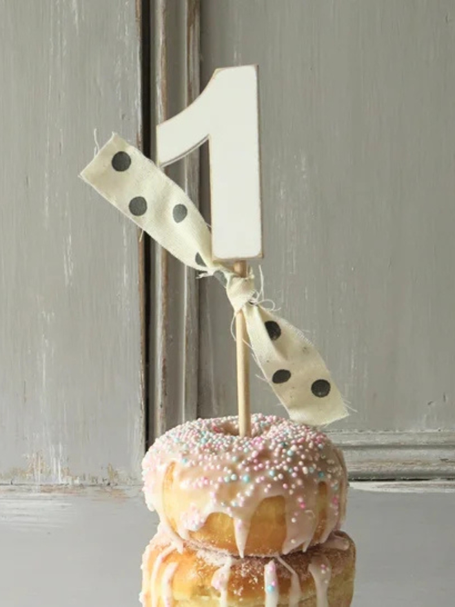 hand painted wooden number cake topper. a white number one cake topper with a spotty ribbon on a cake