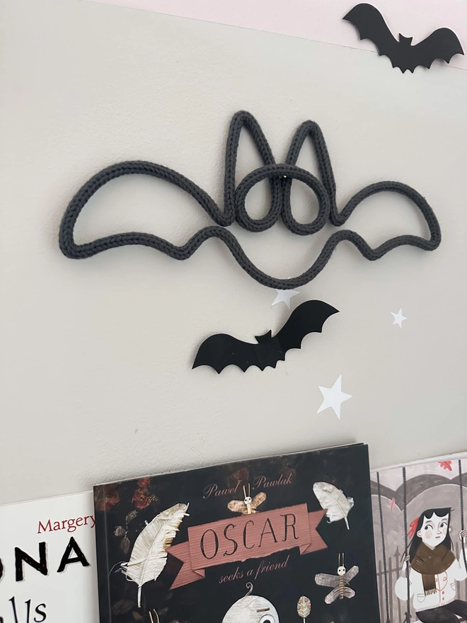 Graphite Colour Knitted Bat Halloween Decor hanging on a brass pin on the wall above some kids halloween books, styled alongsideblack paper bats and star decorations for the wall.