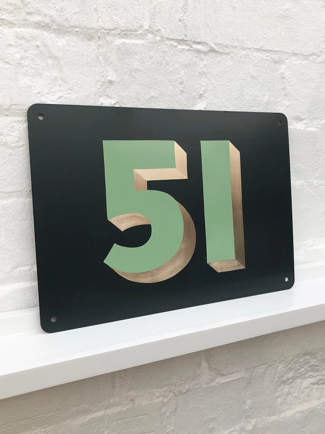 Hand painted sage green and gold leaf house number 51 on an anthracite grey metal plaque against a white brick wall. 