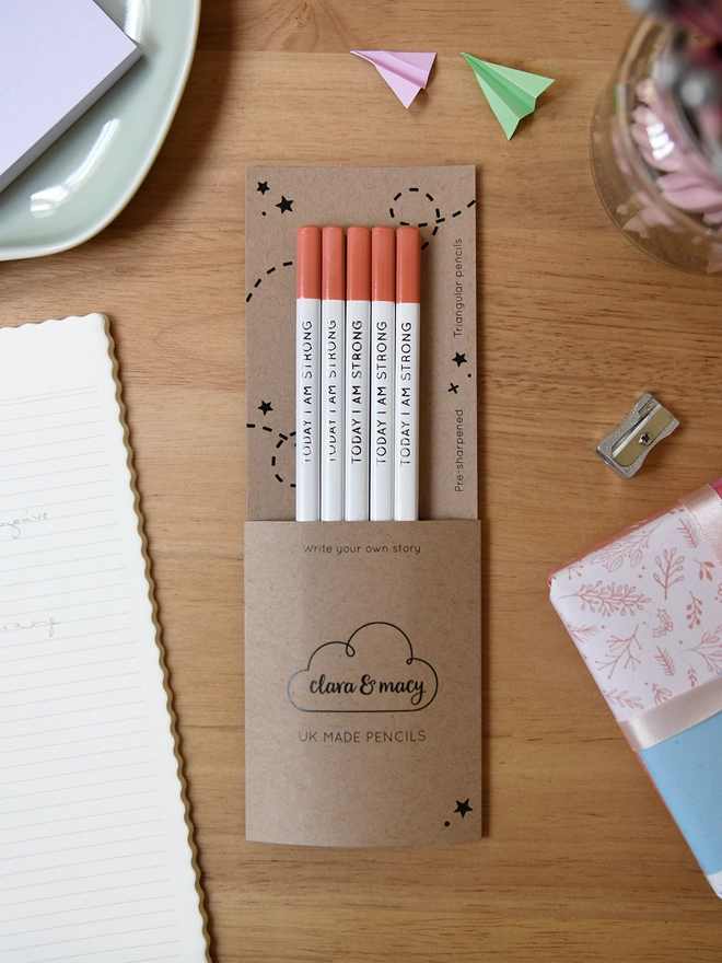Five white pencils with orange tips and the words Today I Am Strong are packed in a cardboard sleeve. It rests on a wooden desk with stationery items around it.