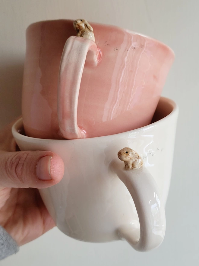 hand holding 2 pottery cups the pink cup is stacked on the white mug and both have a tiny beige bunny rabbit sitting on the top of the handle with tiny paw prints on the cup 