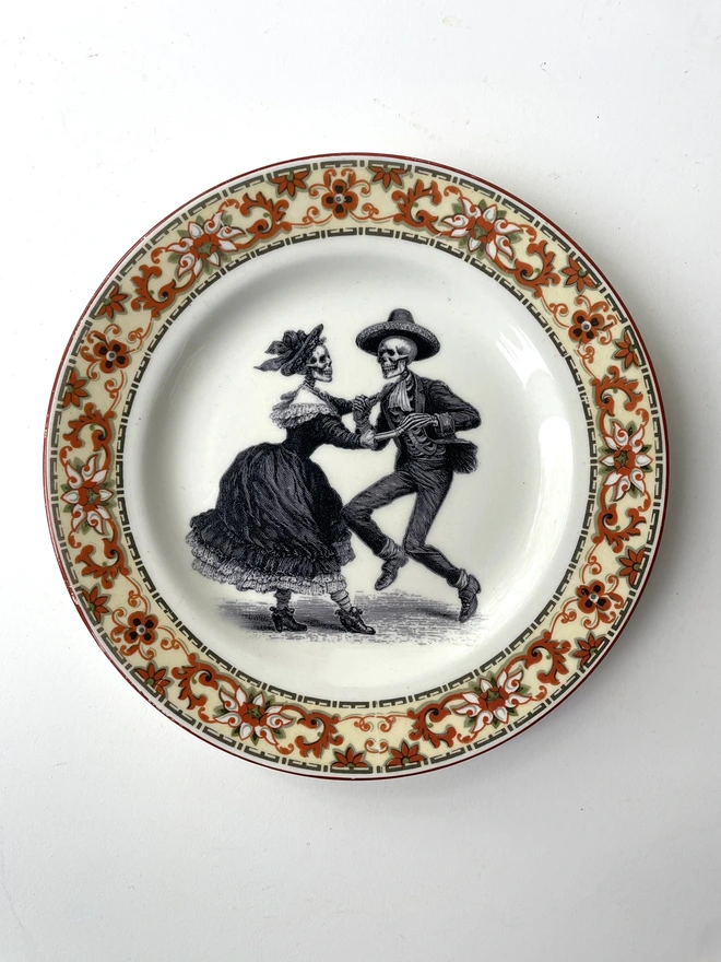 vintage plate with an ornate border, with a printed vintage illustration of two dancing day of the dead skeletons wearing a dress and a suit in the middle