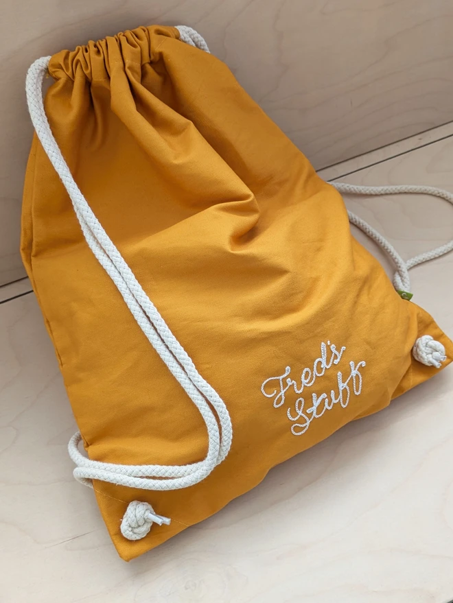 Mustard Drawstring Gymsac Bag sitting casually on a wooden bench, reading 'Fred's Stuff' in white embroidery thread