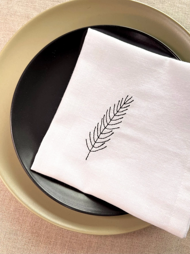White cotton Christmas napkin with embroidered fir leaf design, styled on green and black plates