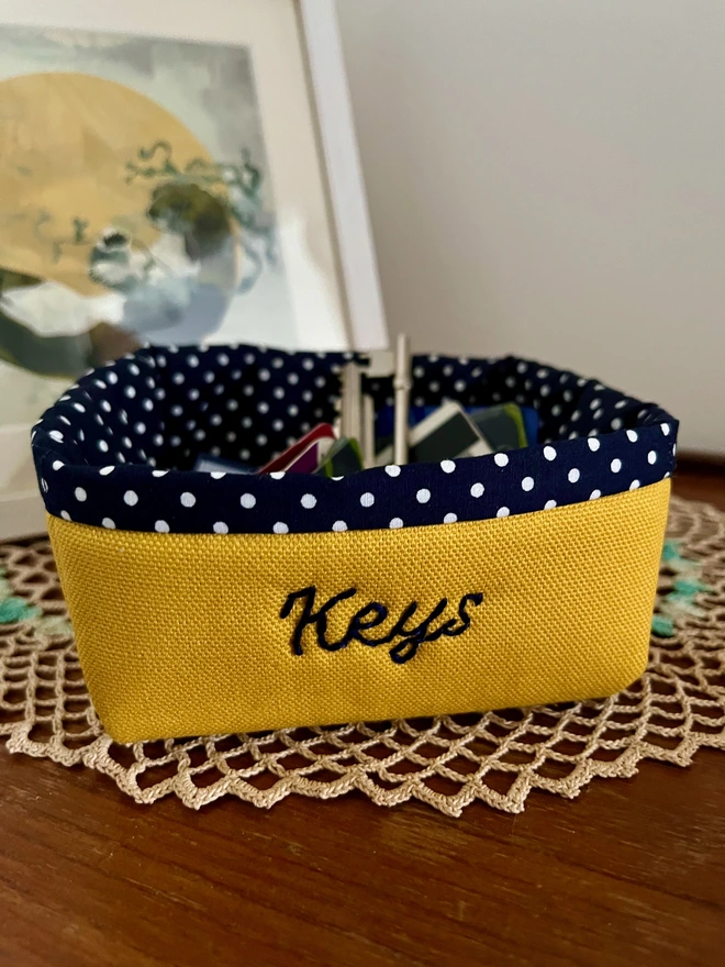 Yellow fabric storage basket with navy polka dot lining, embroidered with ‘Keys’ & holding keys.