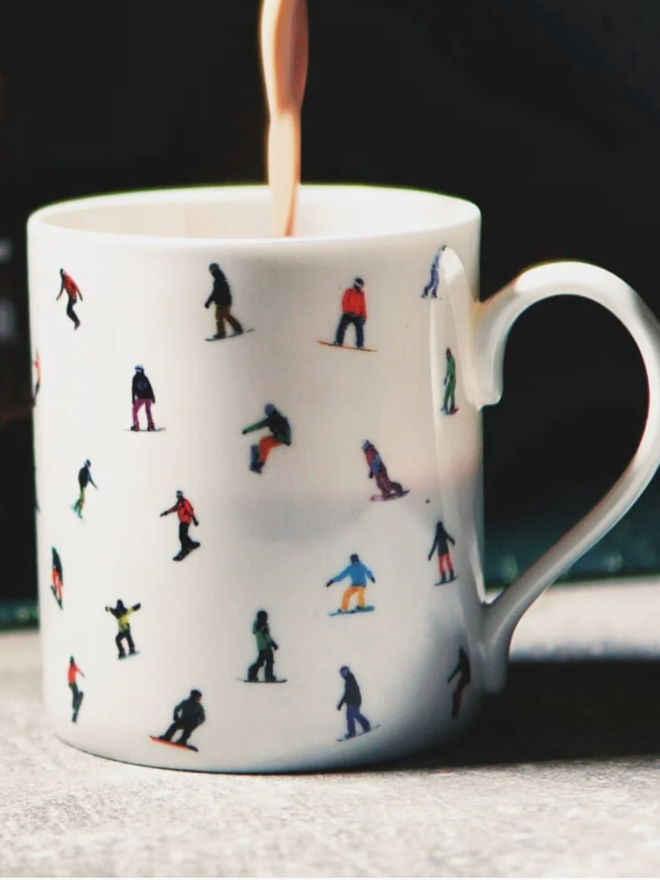 Snowboard Mug. A simple, white mug with a multicoloured pattern of snowboarders. The mug is shown in use with a hot drink being poured into it.