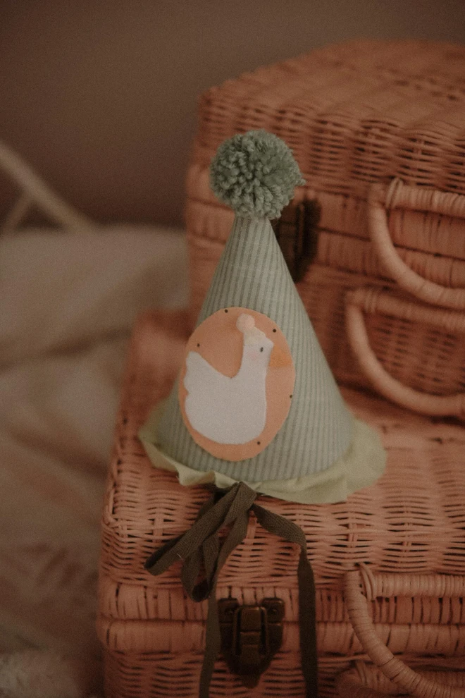 Baby Birthday Hat With Goose, a birthday party hat with a goose. It sits on a wooden hamper. 