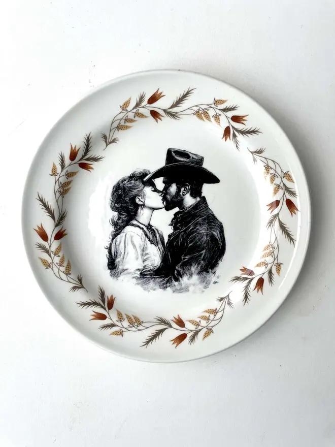 a vintage plate with an ornate border has a black and white vintage illustration of a cowboy and cowgirl sharing a kiss in the centre