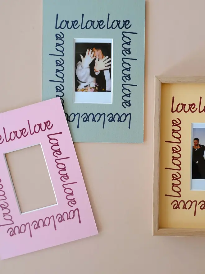 'Love' instax mini mount frame with a blue, purple and yellow mount featuring a decorative frill border around a centered Instax Square photo, displayed on a neutral beige wall.