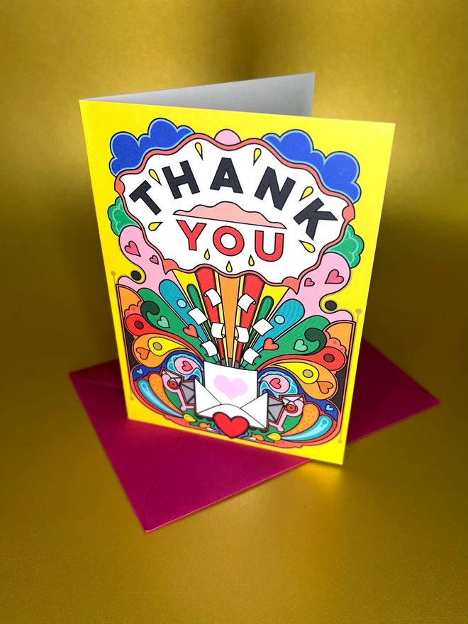 A vibrant yellow card with Thank You written boldly at the top, above a multi-coloured abstract design featuring hearts, envelopes and paper, sits on top of a red envelope on a gold backdrop.