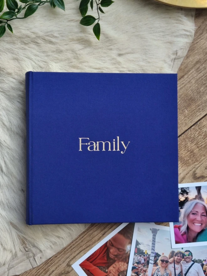 An indigo blue hand-bound photo album printed with the word 'Family' in gold lettering rests on a floor next to family photographs.