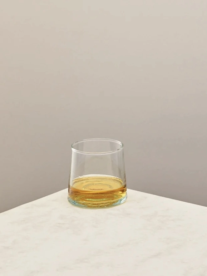Low Glasses (Set Of 1 Or 4), a low glass sitting on a white counter top against a plain backdrop. It is filled by an alcoholic drink. 