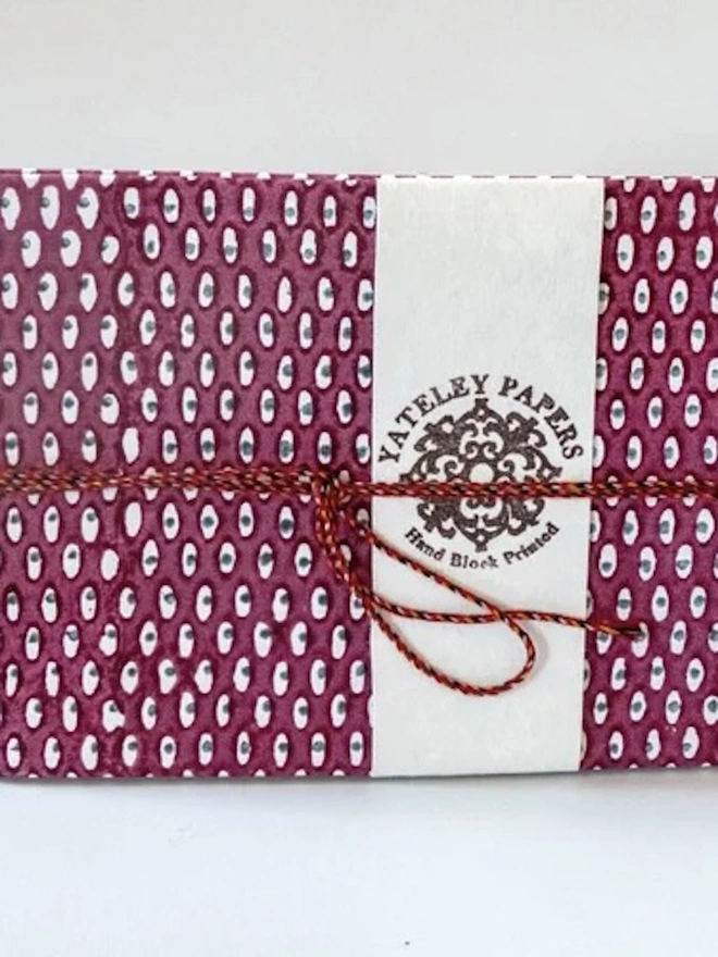 A5 Small Landscape Travel Watercolour Book. A burgundy and white spotted Yateley Papers notebook wrapped in a cream hand block printed paper band and tied with red braided cord. The textured printed cover and simple bow make it a ready-to-give handmade stationery gift.
